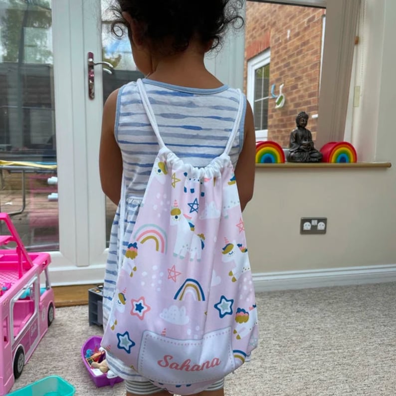 May include: A pink drawstring bag with a unicorn and rainbow print. The bag has a white pocket on the front with the name "Sahana" written in black.