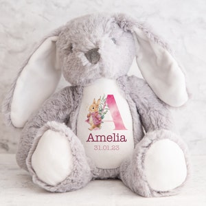 May include: A gray plush bunny with a white patch on its chest. The patch features a pink watercolor letter A with floral accents and the name Amelia and the date 31.01.23.