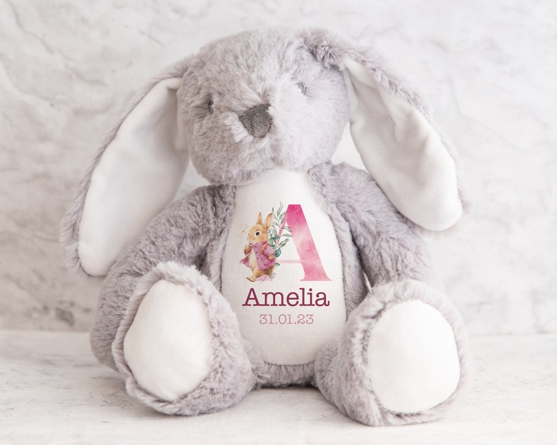 May include: A gray plush bunny with a white patch on its chest. The patch features a pink watercolor letter A with floral accents and the name Amelia and the date 31.01.23.