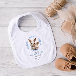 Peut inclure: Bavoir de b&eacute;b&eacute; blanc avec un motif de lapin bleu et blanc. Le bavoir dit "Notre premi&egrave;re f&ecirc;te des p&egrave;res Papa & Jessica".