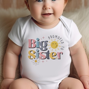 May include: A white baby onesie with the text "Promoted to Big Sister" in colorful letters with a smiling sun and daisies.