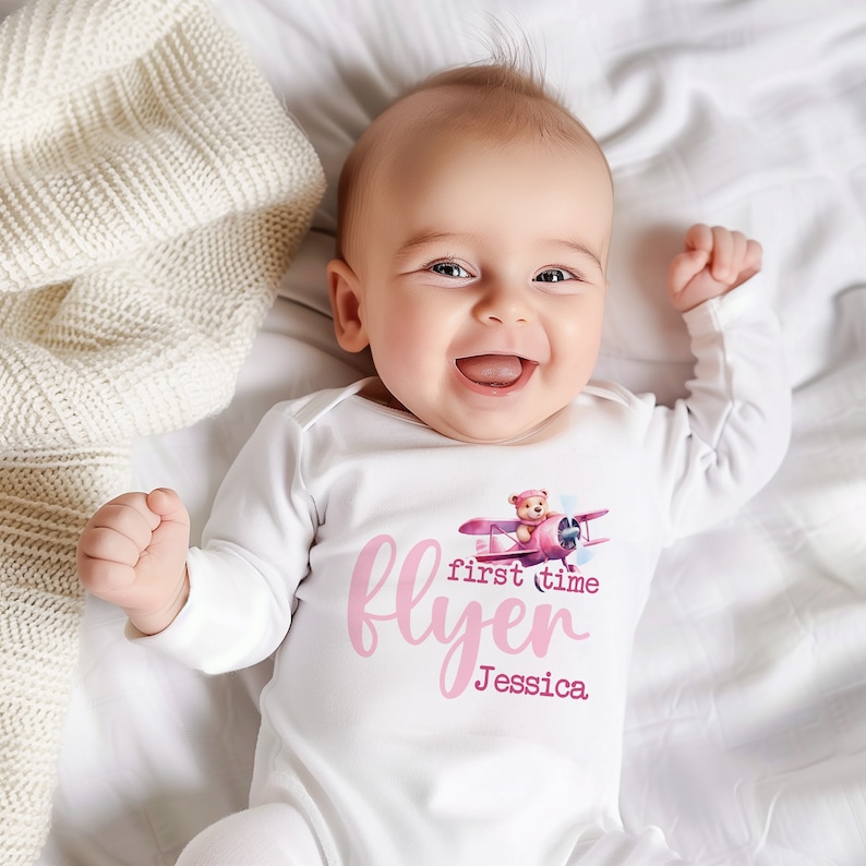 May include: A white baby onesie with a pink graphic of a teddy bear flying a pink airplane. The text "first time flyer Jessica" is printed in pink on the onesie.