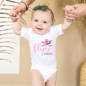 May include: A white baby onesie with a pink airplane and teddy bear graphic. The text "first time flyer Jessica" is printed on the onesie.