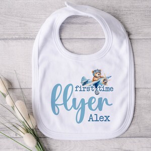 Pu&ograve; includere: Bavaglino bianco per neonato con un design di aereo blu e il testo "first time flyer Alex".