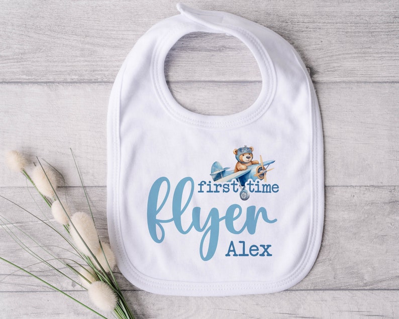 Pu&ograve; includere: Bavaglino bianco per neonato con un design di aereo blu e il testo "first time flyer Alex".