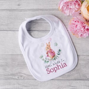 K&ouml;nnte beinhalten: Wei&szlig;er Baby-L&auml;tzchen mit einer Aquarell-Illustration eines Kaninchens, das Ballons h&auml;lt. Der Text "Hello World I'm Sophia" ist in Rosa unter dem Kaninchen gedruckt.
