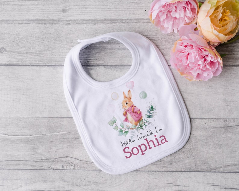 K&ouml;nnte beinhalten: Wei&szlig;er Baby-L&auml;tzchen mit einer Aquarell-Illustration eines Kaninchens, das Ballons h&auml;lt. Der Text "Hello World I'm Sophia" ist in Rosa unter dem Kaninchen gedruckt.