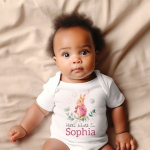 K&ouml;nnte beinhalten: Wei&szlig;er Baby-Strampler mit einem rosa und braunen Kaninchen, das Ballons h&auml;lt. Der Strampler hat den Text "Hello World I'm Sophia" aufgedruckt.