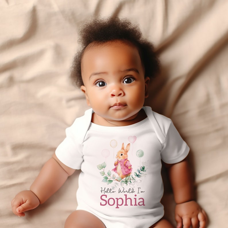 K&ouml;nnte beinhalten: Wei&szlig;er Baby-Strampler mit einem rosa und braunen Kaninchen, das Ballons h&auml;lt. Der Strampler hat den Text "Hello World I'm Sophia" aufgedruckt.