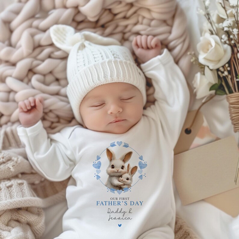 Peut inclure: Une grenouill&egrave;re blanche pour b&eacute;b&eacute; avec des accents bleus repr&eacute;sente deux lapins et le texte "Notre premi&egrave;re f&ecirc;te des p&egrave;res Papa & Jessica".
