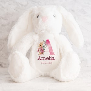 May include: White plush bunny with a pink watercolor letter A and floral design. The bunny has a personalized name "Amelia" and date "31.01.23" printed on the chest.