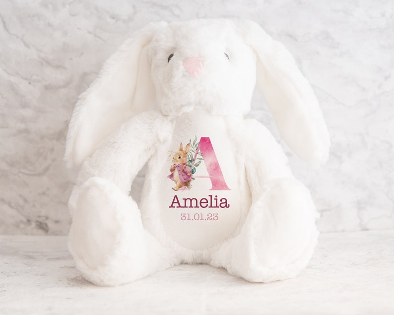 May include: White plush bunny with a pink watercolor letter A and floral design. The bunny has a personalized name "Amelia" and date "31.01.23" printed on the chest.