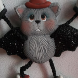 May include: A whimsical, gray bat-like figurine with a red hat, rosy cheeks, and large eyes. It has black glittery wings and is wearing white sneakers with red accents. The figurine is a decorative item.