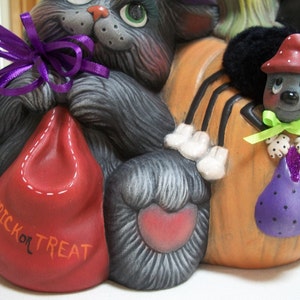 Puede incluir: Decoraci&oacute;n de cer&aacute;mica con tem&aacute;tica de Halloween con un gato gris, una calabaza con una ara&ntilde;a y dos bolsas de golosinas. La bolsa roja dice "TRICK OR TREAT". La ara&ntilde;a lleva un sombrero rojo y sostiene una bolsa morada. El gato tiene una cinta morada.