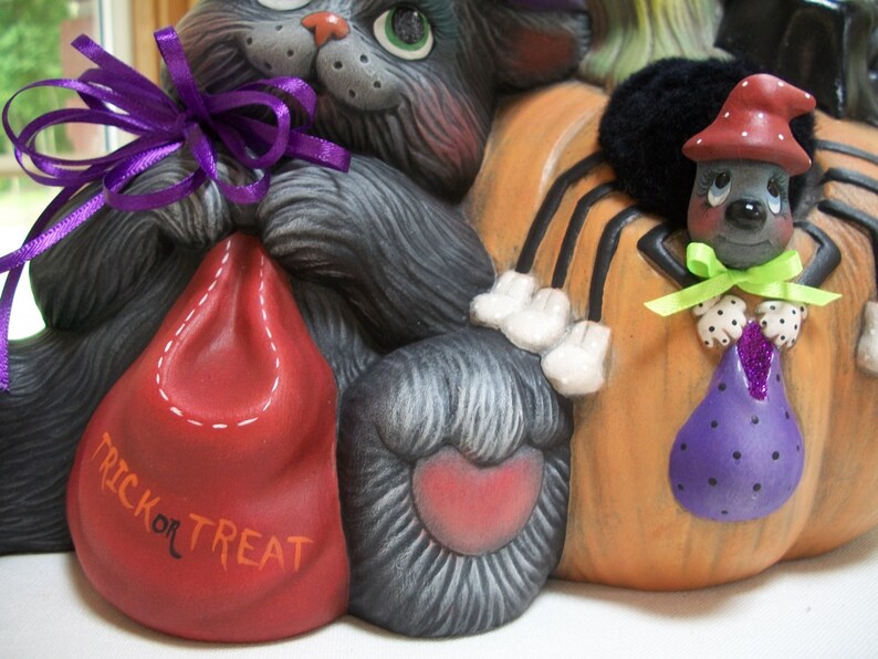 Puede incluir: Decoraci&oacute;n de cer&aacute;mica con tem&aacute;tica de Halloween con un gato gris, una calabaza con una ara&ntilde;a y dos bolsas de golosinas. La bolsa roja dice "TRICK OR TREAT". La ara&ntilde;a lleva un sombrero rojo y sostiene una bolsa morada. El gato tiene una cinta morada.