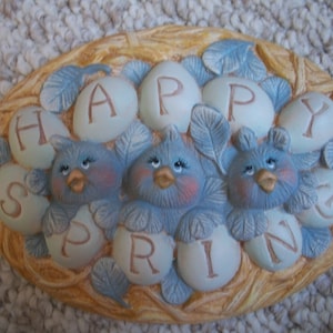 May include: A ceramic wall decor featuring a nest with blue birds and eggs. The eggs are decorated with the word "HAPPY SPRING".