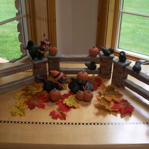 May include: A wooden fence with black cat figurines holding pumpkins and birds. The fence is decorated with fall leaves.