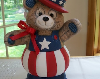 May include: A patriotic teddy bear figurine wearing a red, white, and blue hat with stars. The bear is dressed in a red, white, and blue striped outfit with a white star on the chest. The bear is standing on a white surface.