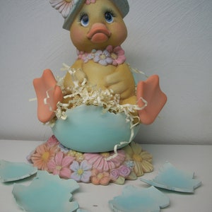 May include: A ceramic duck figurine wearing a blue and white hat with pink flowers. The duck is sitting in a blue egg shell with pink and white flowers on the base. The egg shell is broken into pieces.