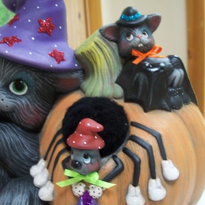 Puede incluir: Decoraci&oacute;n de Halloween de cer&aacute;mica con un gato gris con un sombrero de bruja morado con estrellas de purpurina roja, un murci&eacute;lago negro y una ara&ntilde;a sobre una calabaza naranja. El gato tiene ojos verdes y sostiene una bolsa morada.