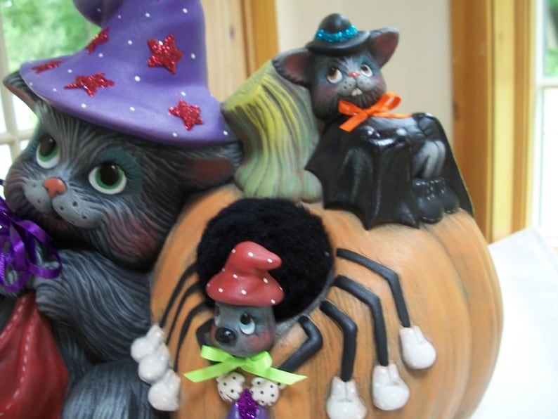 Puede incluir: Decoraci&oacute;n de Halloween de cer&aacute;mica con un gato gris con un sombrero de bruja morado con estrellas de purpurina roja, un murci&eacute;lago negro y una ara&ntilde;a sobre una calabaza naranja. El gato tiene ojos verdes y sostiene una bolsa morada.