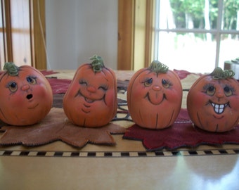 May include: Four hand-painted ceramic pumpkins with different facial expressions. The pumpkins are orange with green stems and painted details. They are arranged on a red and brown patterned surface.