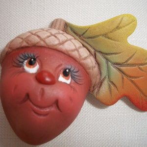 May include: A ceramic acorn figurine with a smiling face, eyelashes, and a small red nose. The acorn has a brown cap and is attached to a colourful leaf with yellow, green, and orange hues. The figurine is likely a fall or autumn decoration.