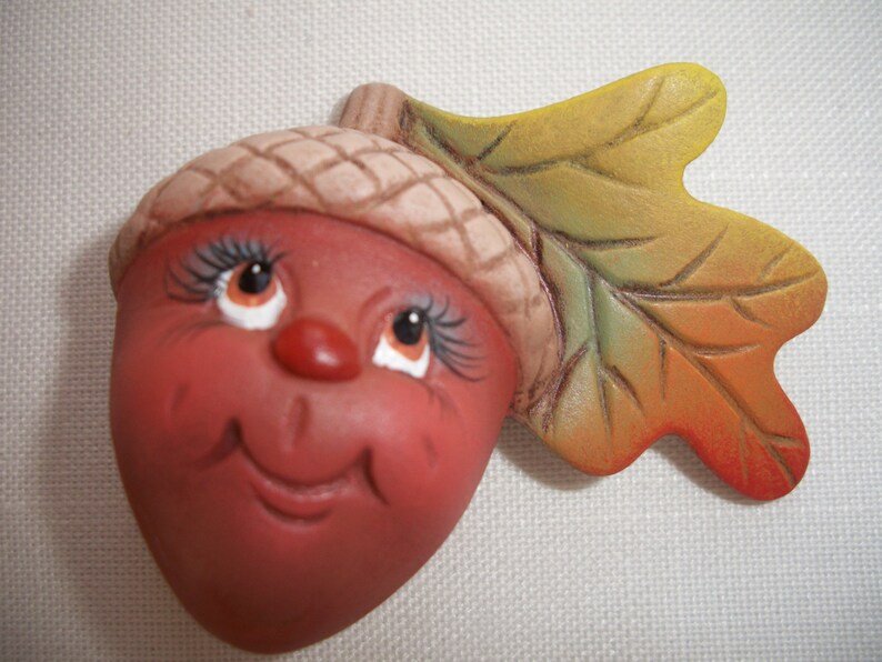 May include: A ceramic acorn figurine with a smiling face, eyelashes, and a small red nose. The acorn has a brown cap and is attached to a colourful leaf with yellow, green, and orange hues. The figurine is likely a fall or autumn decoration.