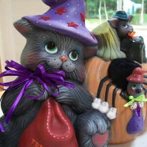 Puede incluir: Decoraci&oacute;n de Halloween de cer&aacute;mica. Un gato gris lleva un sombrero de bruja morado con estrellas de purpurina roja. El gato sostiene una bolsa roja con "TRICK". Una calabaza con una ara&ntilde;a negra y dos gatos negros completa la escena.