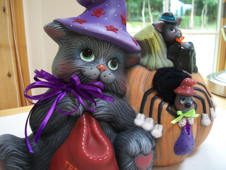 Puede incluir: Decoraci&oacute;n de Halloween de cer&aacute;mica. Un gato gris lleva un sombrero de bruja morado con estrellas de purpurina roja. El gato sostiene una bolsa roja con "TRICK". Una calabaza con una ara&ntilde;a negra y dos gatos negros completa la escena.