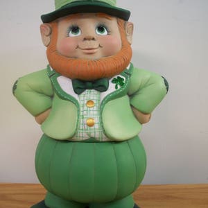 May include: A green and white ceramic figurine of a leprechaun wearing a green hat with a shamrock on top. He has a red beard and is wearing a green jacket with gold buttons and a green bow tie.