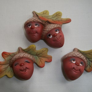May include: Three ceramic acorn figurines with smiling faces and attached leaves. The acorns are painted in shades of brown and red, with green and yellow leaves. The faces have detailed eyes and rosy cheeks.
