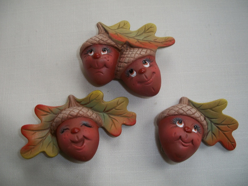 May include: Three ceramic acorn figurines with smiling faces and attached leaves. The acorns are painted in shades of brown and red, with green and yellow leaves. The faces have detailed eyes and rosy cheeks.