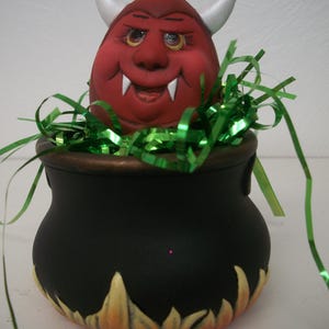 May include: A ceramic Halloween decoration featuring a red devil head with white horns and fangs emerging from a black cauldron. The cauldron has yellow and orange flames painted around the base, and green decorative ribbon is inside.