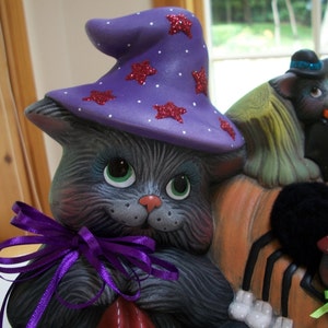 Puede incluir: Decoraci&oacute;n de Halloween de cer&aacute;mica con un gato gris que lleva un sombrero de bruja morado con estrellas rojas brillantes. El gato tiene ojos verdes y una cinta morada. Otras figuras incluyen un gato negro y una ara&ntilde;a.
