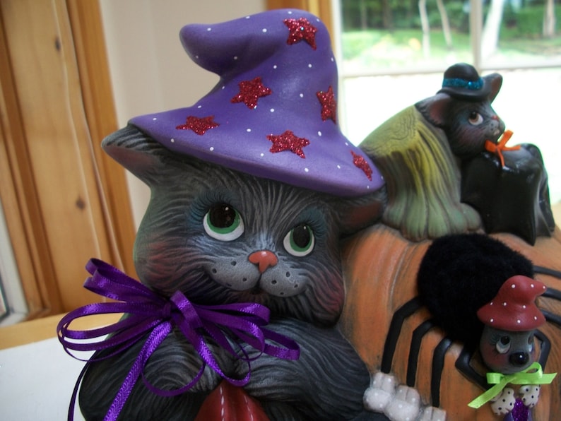 Puede incluir: Decoraci&oacute;n de Halloween de cer&aacute;mica con un gato gris que lleva un sombrero de bruja morado con estrellas rojas brillantes. El gato tiene ojos verdes y una cinta morada. Otras figuras incluyen un gato negro y una ara&ntilde;a.