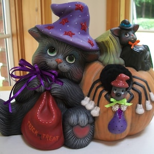 Puede incluir: Decoraci&oacute;n de Halloween de cer&aacute;mica con un gato gris con un sombrero de bruja morado con estrellas rojas, sosteniendo una bolsa roja que dice "TRICK OR TREAT". Un gato negro y una ara&ntilde;a tambi&eacute;n forman parte del dise&ntilde;o, todo sobre una calabaza.