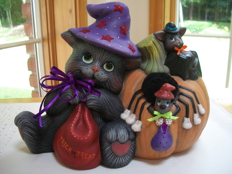 Puede incluir: Decoraci&oacute;n de Halloween de cer&aacute;mica con un gato gris con un sombrero de bruja morado con estrellas rojas, sosteniendo una bolsa roja que dice "TRICK OR TREAT". Un gato negro y una ara&ntilde;a tambi&eacute;n forman parte del dise&ntilde;o, todo sobre una calabaza.