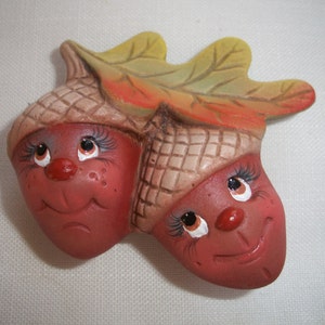 May include: A charming ceramic ornament depicting two smiling acorn characters. The acorns are a reddish-brown colour with tan caps, connected by a leaf with yellow and orange tones. The faces have painted eyes, eyelashes, and red noses.