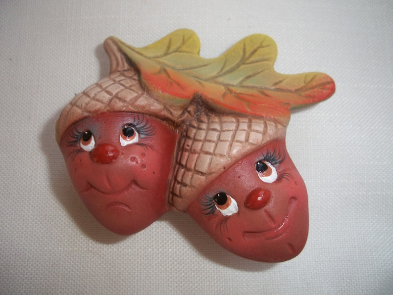 May include: A charming ceramic ornament depicting two smiling acorn characters. The acorns are a reddish-brown colour with tan caps, connected by a leaf with yellow and orange tones. The faces have painted eyes, eyelashes, and red noses.