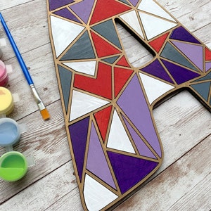 Puede incluir: Una letra A de madera pintada con formas geom&eacute;tricas en tonos de rojo, morado, gris y blanco. La letra est&aacute; rodeada de botes de pintura de varios colores, incluyendo negro, blanco, rosa, amarillo, azul y verde.