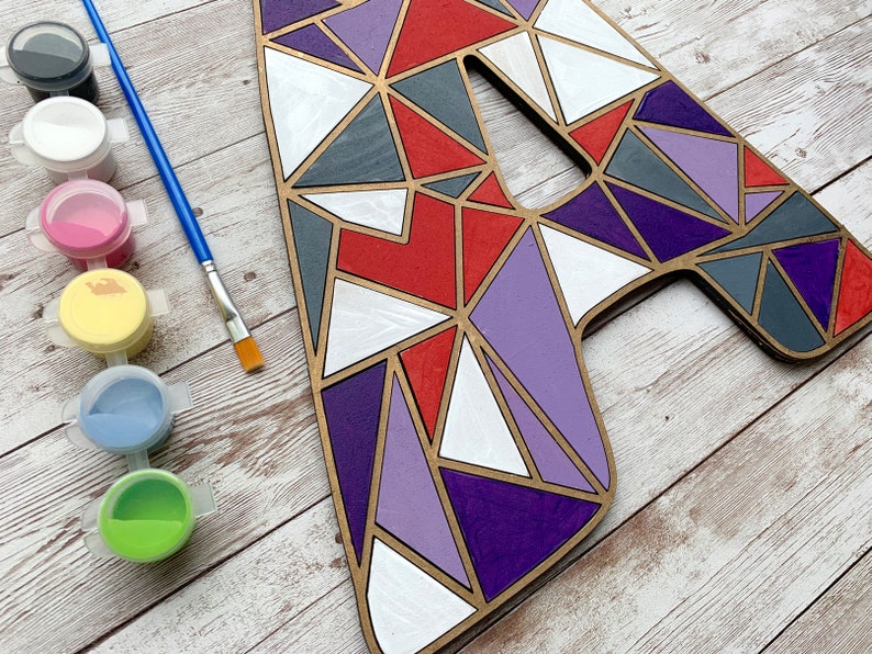 Puede incluir: Una letra A de madera pintada con formas geom&eacute;tricas en tonos de rojo, morado, gris y blanco. La letra est&aacute; rodeada de botes de pintura de varios colores, incluyendo negro, blanco, rosa, amarillo, azul y verde.
