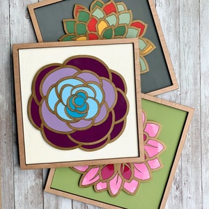 May include: Three framed wood wall art pieces featuring different colored floral designs. The top piece is gray with a red, yellow, and green flower. The middle piece is white with a purple and blue flower. The bottom piece is green with a pink flower.