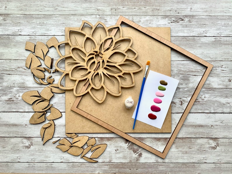 May include: A wooden craft kit featuring a large flower, leaves, and a frame. The kit includes paint colors in shades of green, pink, and red, a paintbrush, and a small bottle of glue. "Glue" is written on the bottle.