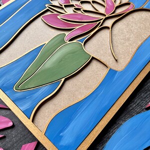 Op de afbeelding: Een houten wanddecoratie met een roze en witte bloem met groene bladeren, tegen een blauwe en gouden achtergrond.