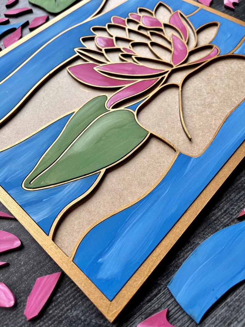 Op de afbeelding: Een houten wanddecoratie met een roze en witte bloem met groene bladeren, tegen een blauwe en gouden achtergrond.