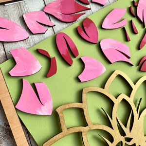 May include: A wooden craft project featuring a large gold mandala with pink and red flower petals surrounding it. The petals are painted and arranged on a green background.