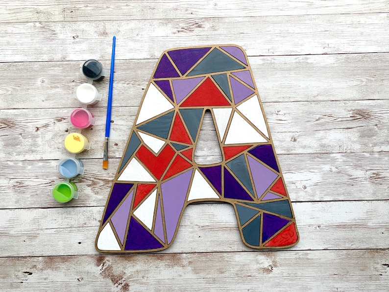 Puede incluir: Una letra de madera A pintada con un patr&oacute;n geom&eacute;trico en tonos de morado, rojo, blanco y gris. La letra est&aacute; rodeada de botes de pintura de varios colores.