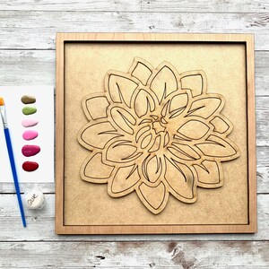 May include: A wooden paint-by-numbers kit featuring a large, detailed outline of a dahlia flower. The kit includes a paintbrush, a small jar of glue, and a palette of six paint colors: green, pink, red, and three shades of pink.