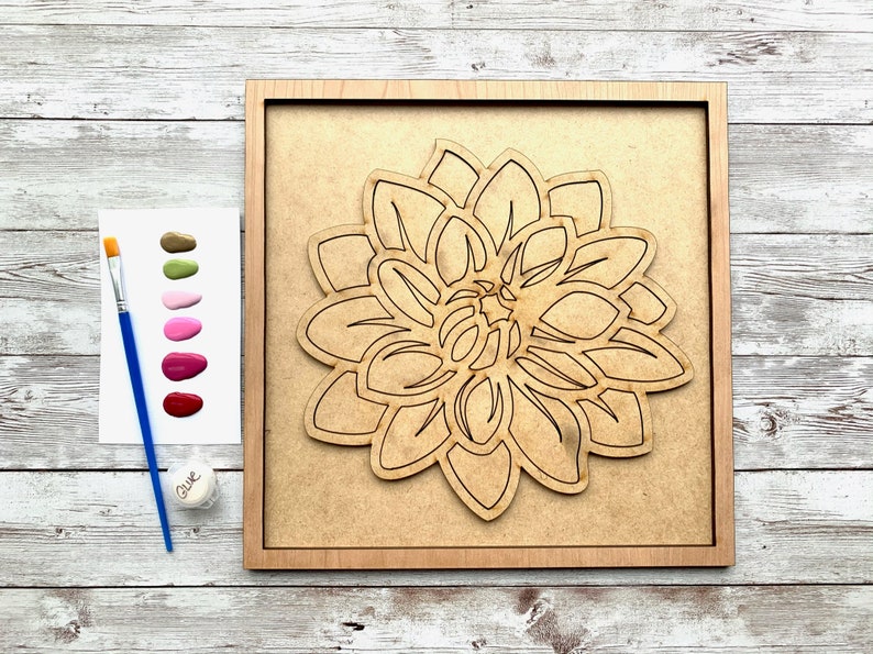 May include: A wooden paint-by-numbers kit featuring a large, detailed outline of a dahlia flower. The kit includes a paintbrush, a small jar of glue, and a palette of six paint colors: green, pink, red, and three shades of pink.